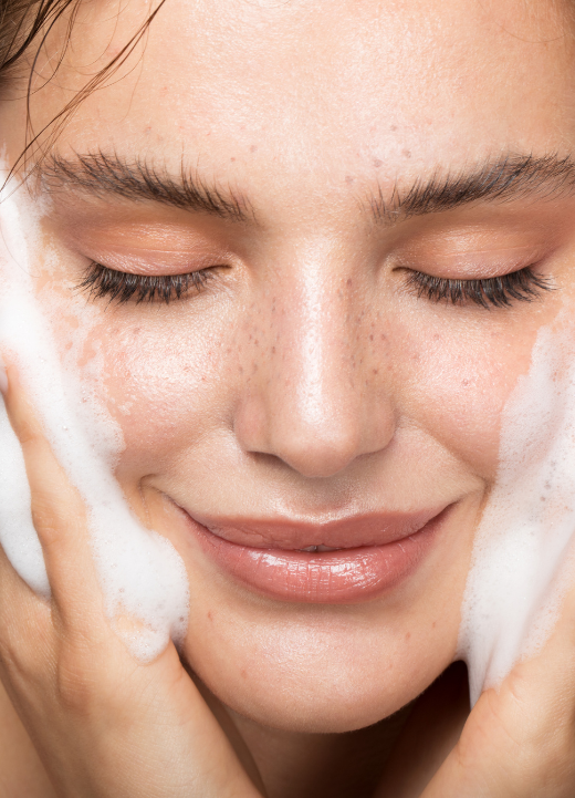 Frontale Nahaufnahme des Gesichts einer Frau, die Reinigungsschaum auf ihren Wangen verteilt.